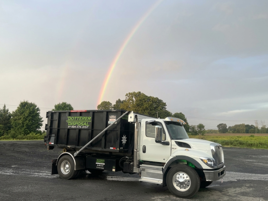 A Dotterer Disposal dumpster truck in a parking lot. A double rainbow is in the sky in the background.