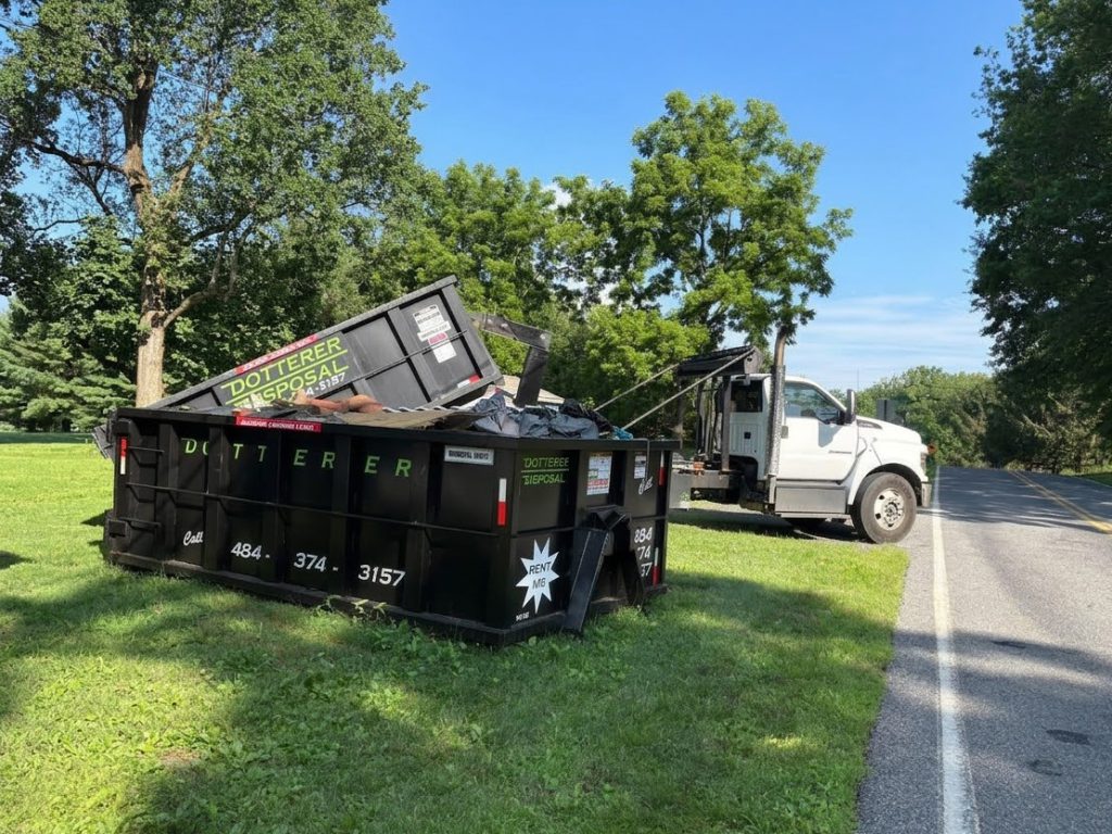 A Dotterer Disposal dumpster filled with junk near a road. A Dotterer Disposal truck is behind it.